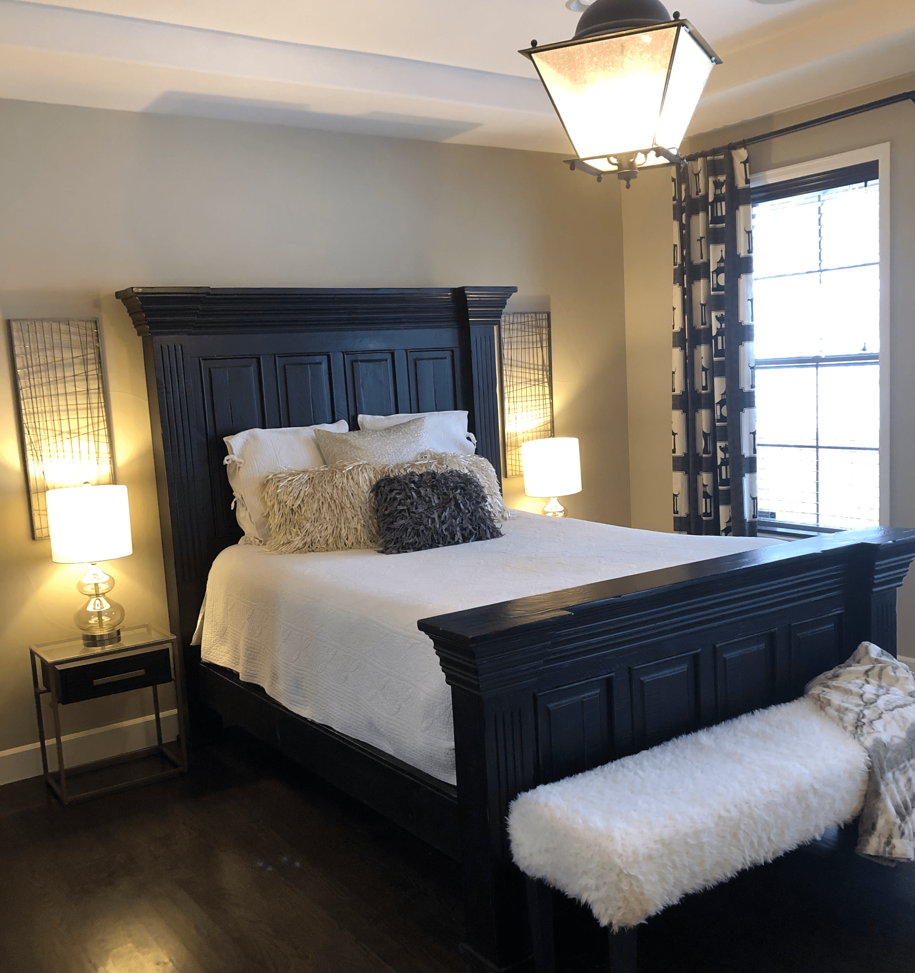 Staged bedroom with a dark carved bed, two lit lamps and a sheepskin bench